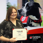 Shannon Noonan displays the posthumous certificate recognizing her dog Blue, the original therapy dog at Carleton, with a photo of Blue behind her.